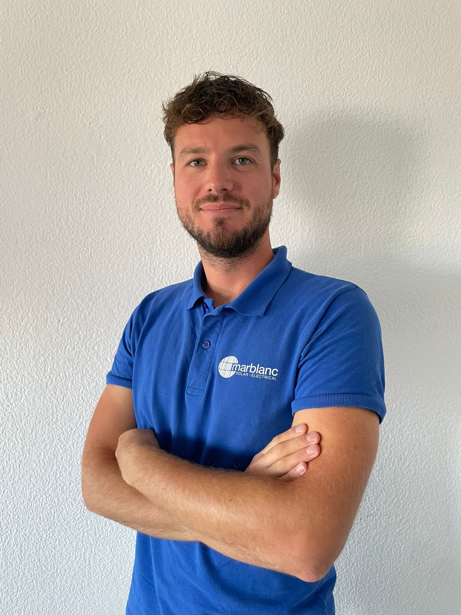 A man with a beard and short hair wearing a blue polo shirt with crossed arms standing against a plain white wall.