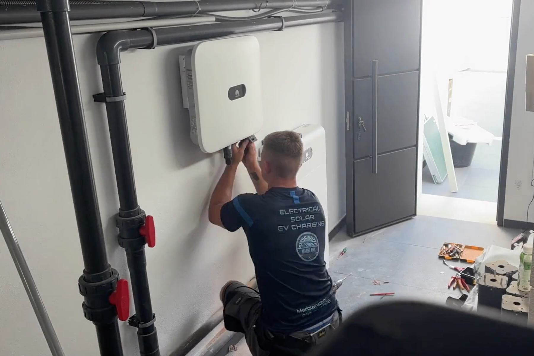 A man crouched down, working on or inspecting a white wall-mounted device with pipes around it in a room.