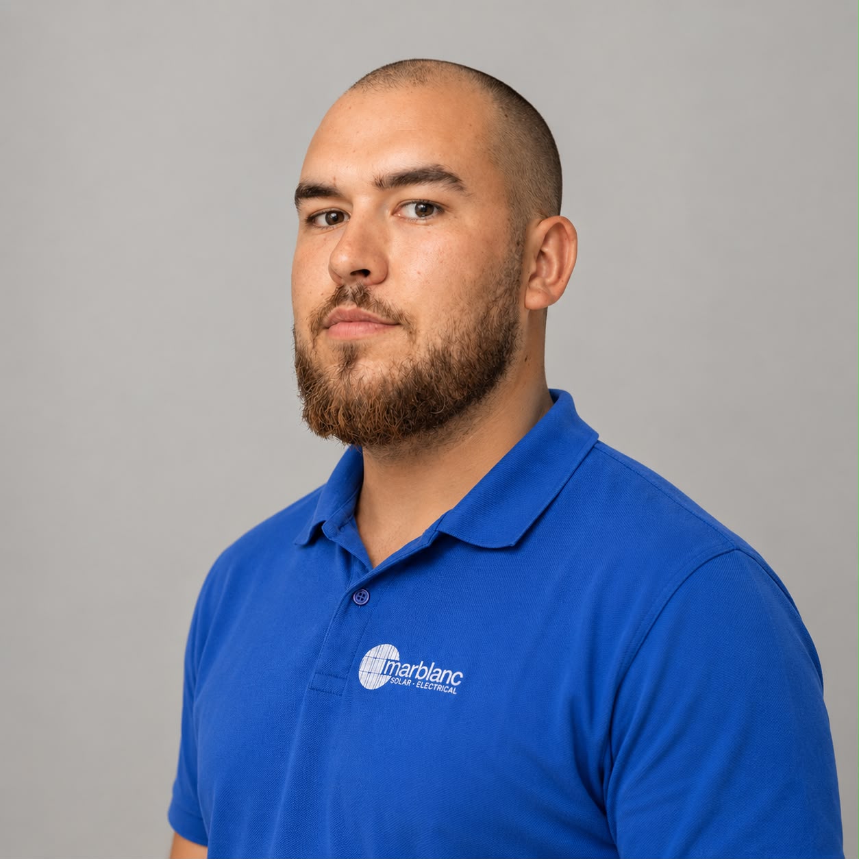 Portrait of a man with a shaved head and beard wearing a blue polo shirt with a company logo, posed against a light gray background, looking toward the camera.