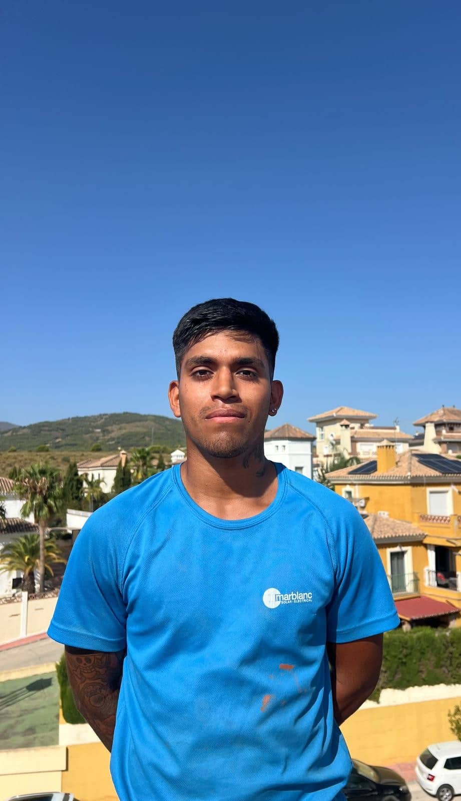 Young man standing outdoors on a sunny day, wearing a blue T‑shirt with a small logo, with houses and mountains in the background